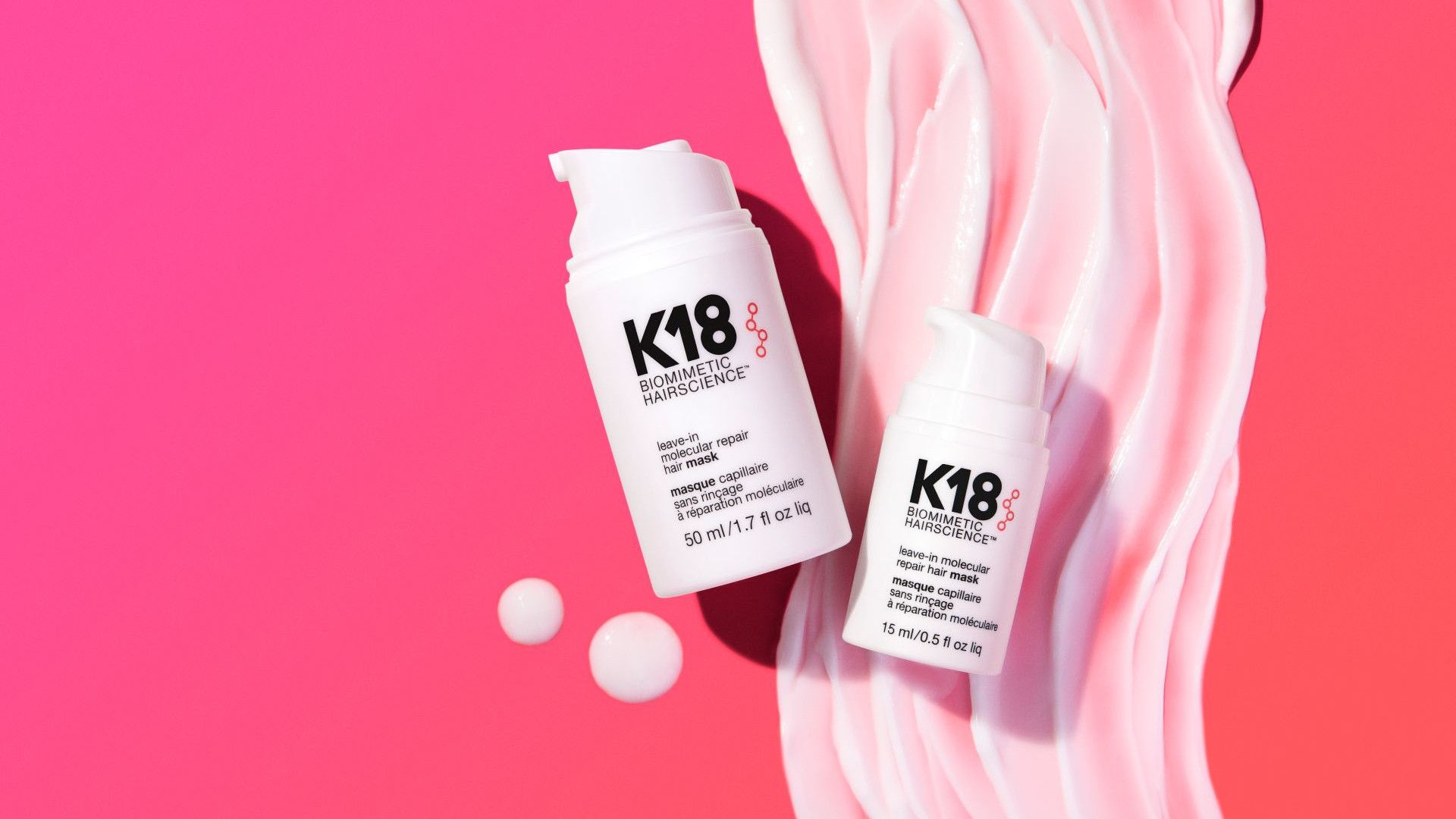 ขวดผลิตภัณฑ์บำรุงผม K18 สีขาว 2 ขวดวางอยู่บนพื้นหลังสีชมพูที่มีลวดลายสีครีมวนเป็นเกลียว
