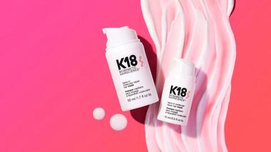 ขวดผลิตภัณฑ์บำรุงผม K18 สีขาว 2 ขวดวางอยู่บนพื้นหลังสีชมพูที่มีลวดลายสีครีมวนเป็นเกลียว