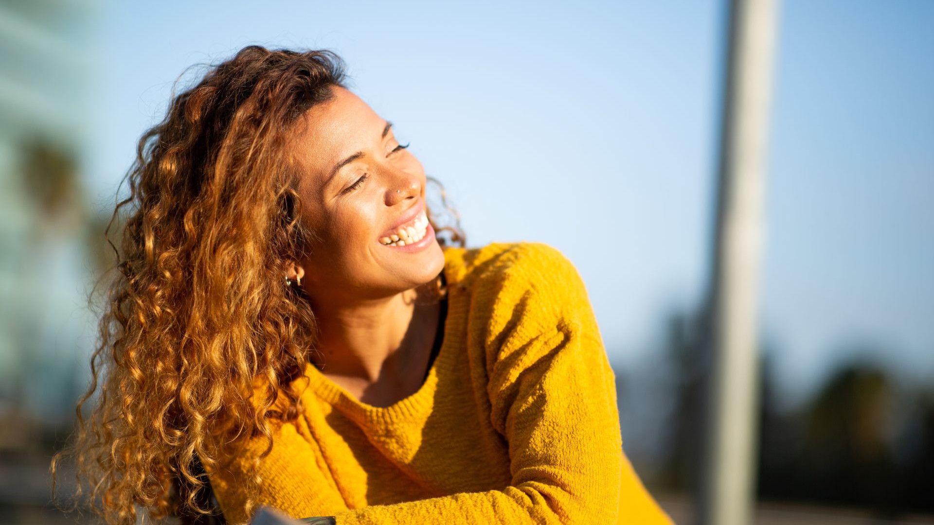 ผู้หญิงสวมเสื้อกันหนาวไหมพรมสีส้ม ยืนอยู่ท่ามกลางทุ่งดอกไม้ใต้ท้องฟ้าสีฟ้าสดใส และมีกำแพงหินแห้งเป็นฉากหลัง